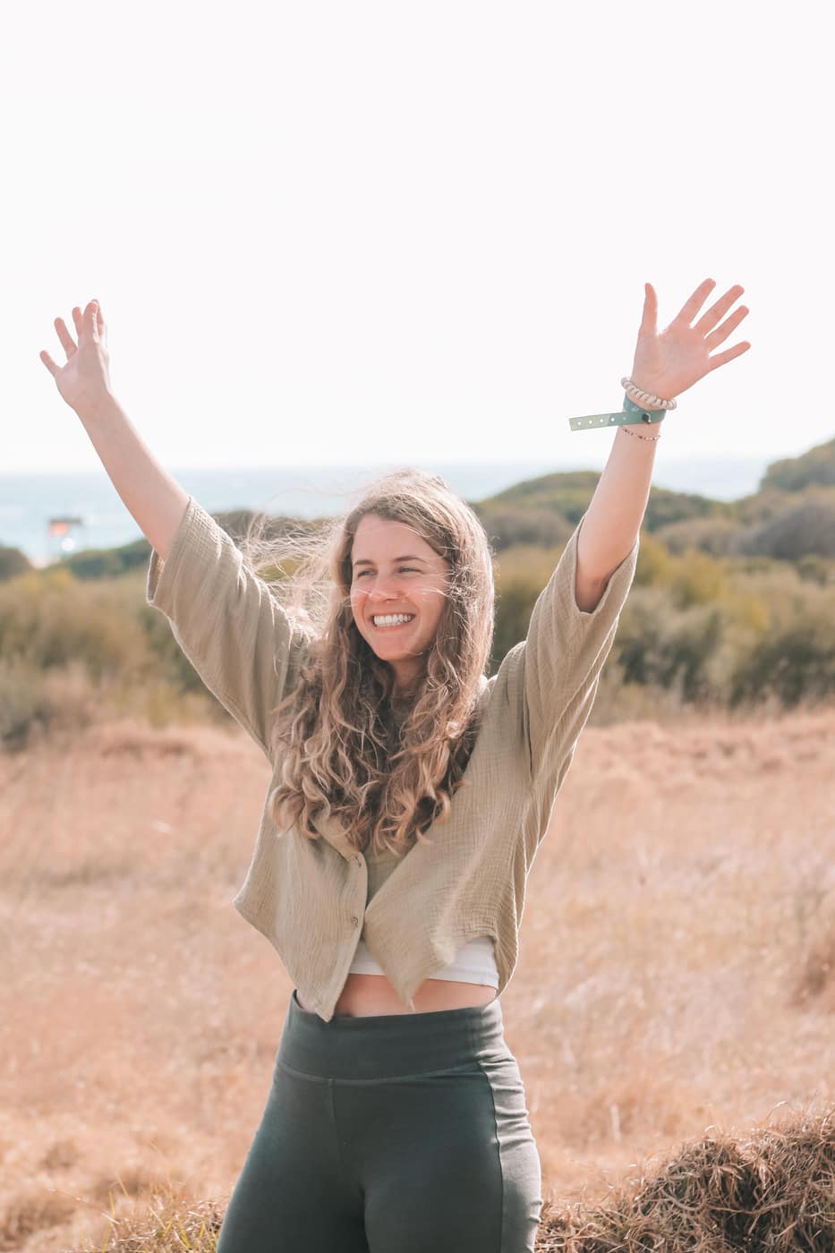Woman smiling with arms raised in a sunny coastal landscape